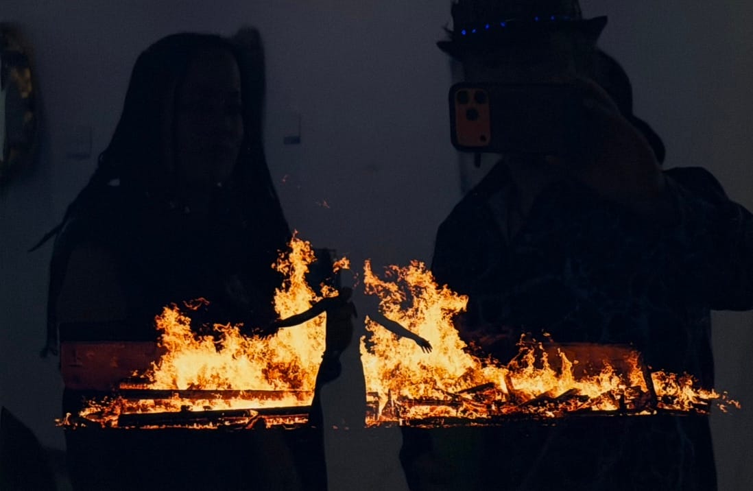 A silhouetted figure surrounded by flames in a reflective artwork exhibited at heARTburn 2026